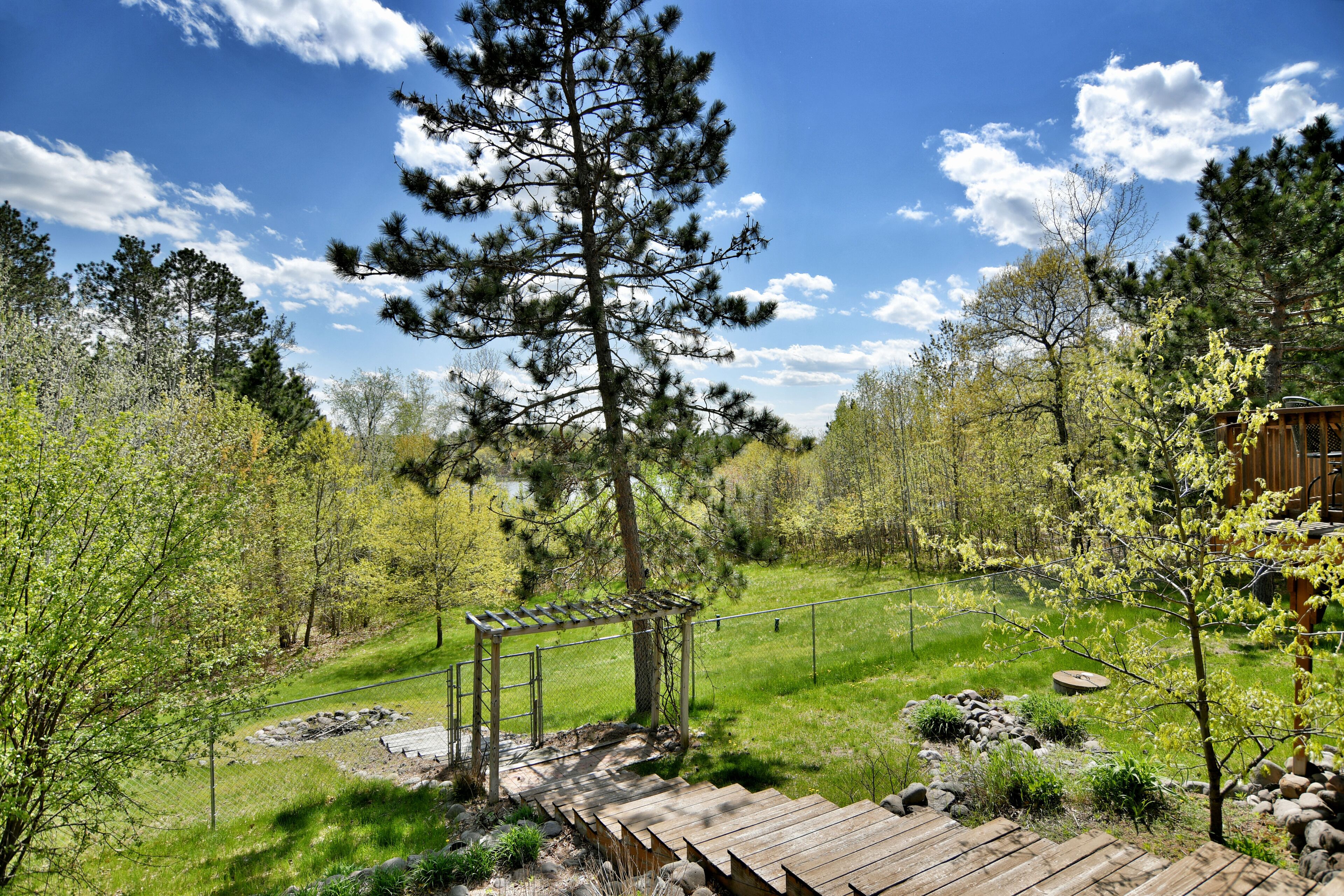 A wooden deck with a pergola, surrounded by a grassy area with trees and a wooden fence.