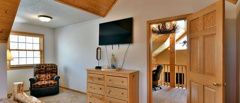 A room with a wooden ceiling, a television mounted on the wall, a wooden dresser, a patterned chair, and a door leading to another room.