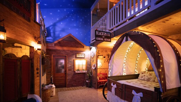 A rustic-themed room with a bed, a sign for Blacksmith and General Repair, and a wooden door labeled "Bank". - Go West- Couples 1-of-a-kind old-time theme-ATL area. (Carroll County)