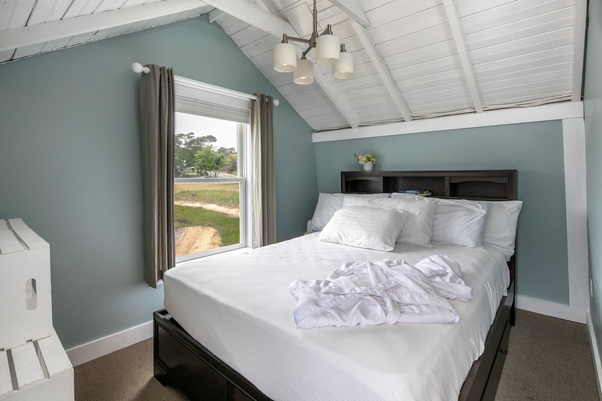 A bedroom with a bed, a window with curtains, a ceiling fan, and a nightstand with a lamp.