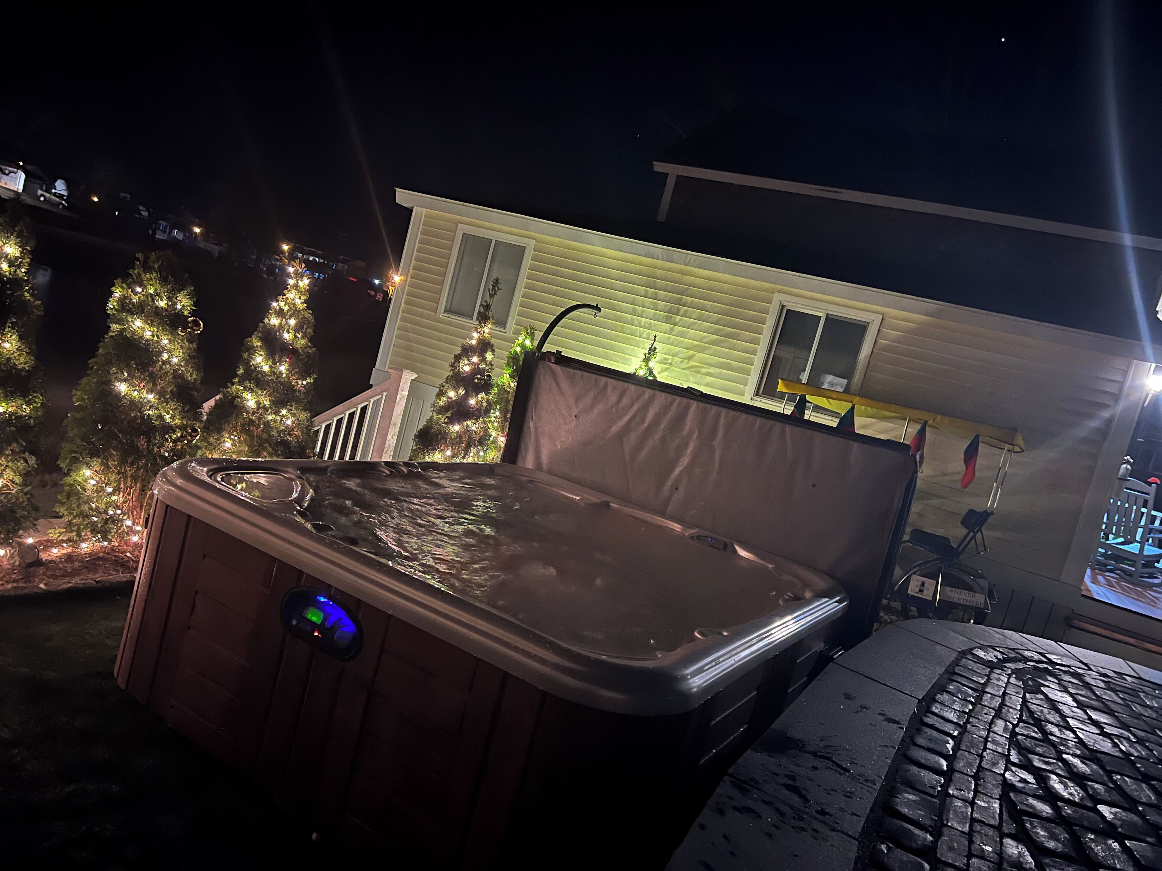 A hot tub with a digital control panel, a covered patio area, and a house with Christmas lights in the background.