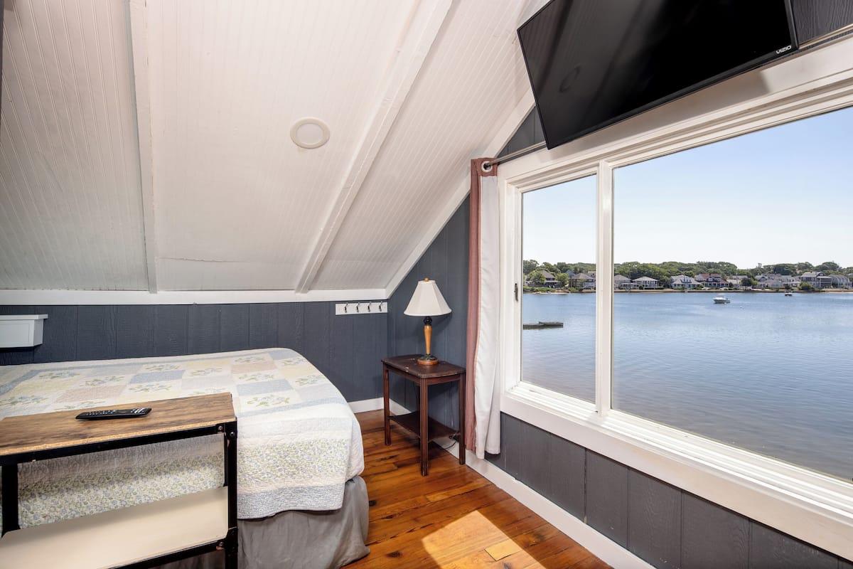 A bedroom with a bed, a small table, a lamp, and a view of a body of water through a large window.