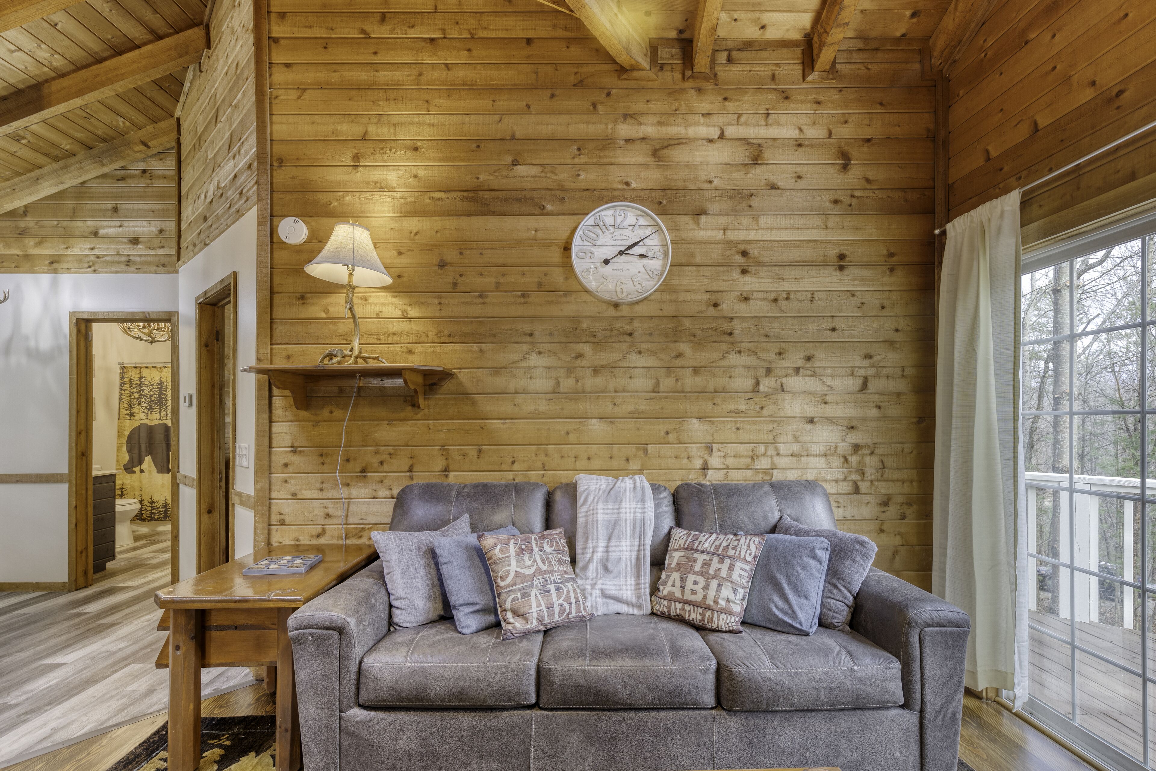 A cozy cabin interior with a wooden ceiling, a gray sofa with patterned cushions, a wall clock, and a window with a view of trees.
