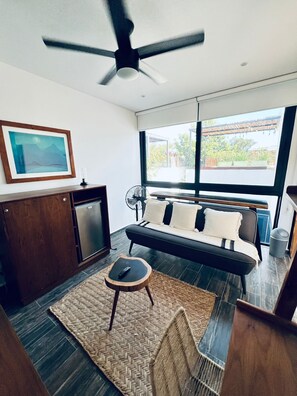 A modern living room with a sofa, a small table, a ceiling fan, a framed picture, and a view of the outdoors.