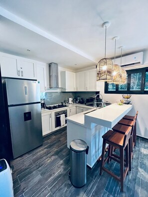 A modern kitchen with white cabinets, stainless steel appliances, a large island with bar stools, and hanging wicker pendant lights.