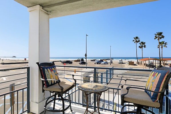Un balcon avec deux chaises et une petite table, donnant sur une plage bordée de palmiers et offrant une vue sur un ciel dégagé.