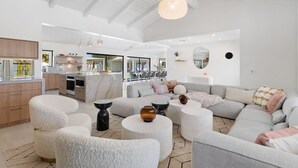 A spacious living area with a large gray sofa, white pouf chairs, and a round coffee table. A kitchen with a marble island and modern appliances is visible in the background. - Solaris - Modern Mediterranean Estate with Pool, Spa and Fire Pit (Palm Springs)