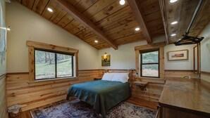 Un dormitorio de cabaña de madera con cama, dos ventanas, una mesita de noche y un escritorio de madera.