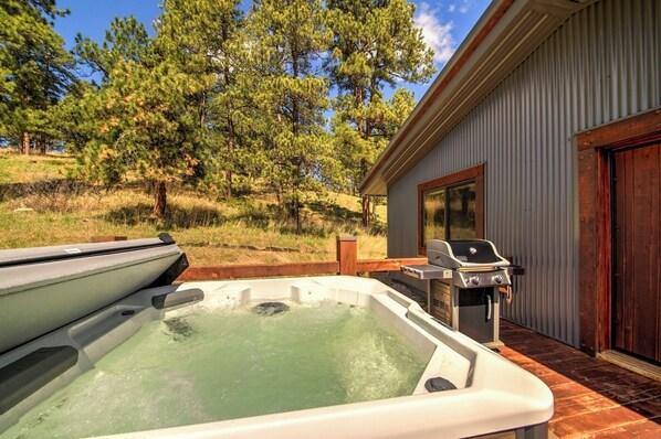 A hot tub with a wooden deck, a barbecue grill, and a building with a large window. - Rustic Custom Timber Frame Cabin (Rapid City)