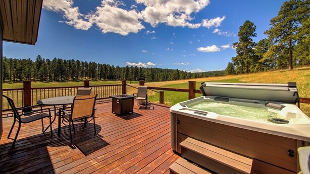 Terraza de madera con mobiliario exterior, un jacuzzi y una vista panorámica de un bosque y un campo abierto.