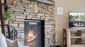 A stone fireplace with a fire, a television on a wooden stand, and a stone wall. - Lake Front, NEW Sandy Beach, Big Buck Hunter & Pac-Man Arcade Games! Near Nisswa (Pequot Lakes)
