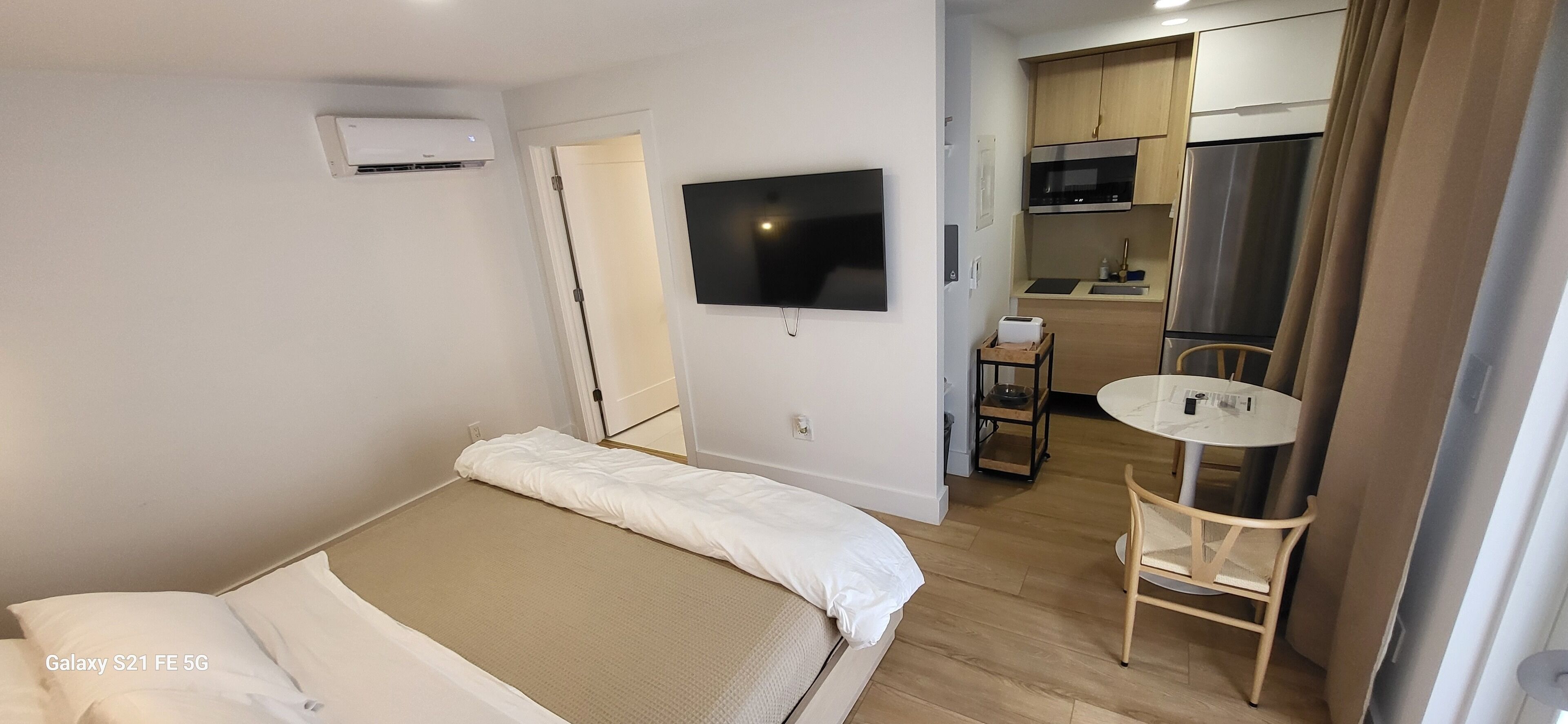 Ein Hotelzimmer mit Bett, Wandfernseher, kleinem Tisch und einer Küchenzeile im Hintergrund.