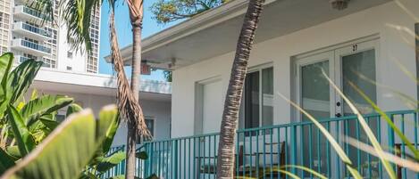 A white building with a balcony, surrounded by palm trees and a white picket fence.