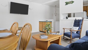 A living room with a TV, wooden dining table, chairs, a blue armchair, and a small kitchen area. - Outpost: Pitchfork 2106 - Walk to Town Square! (Jackson)