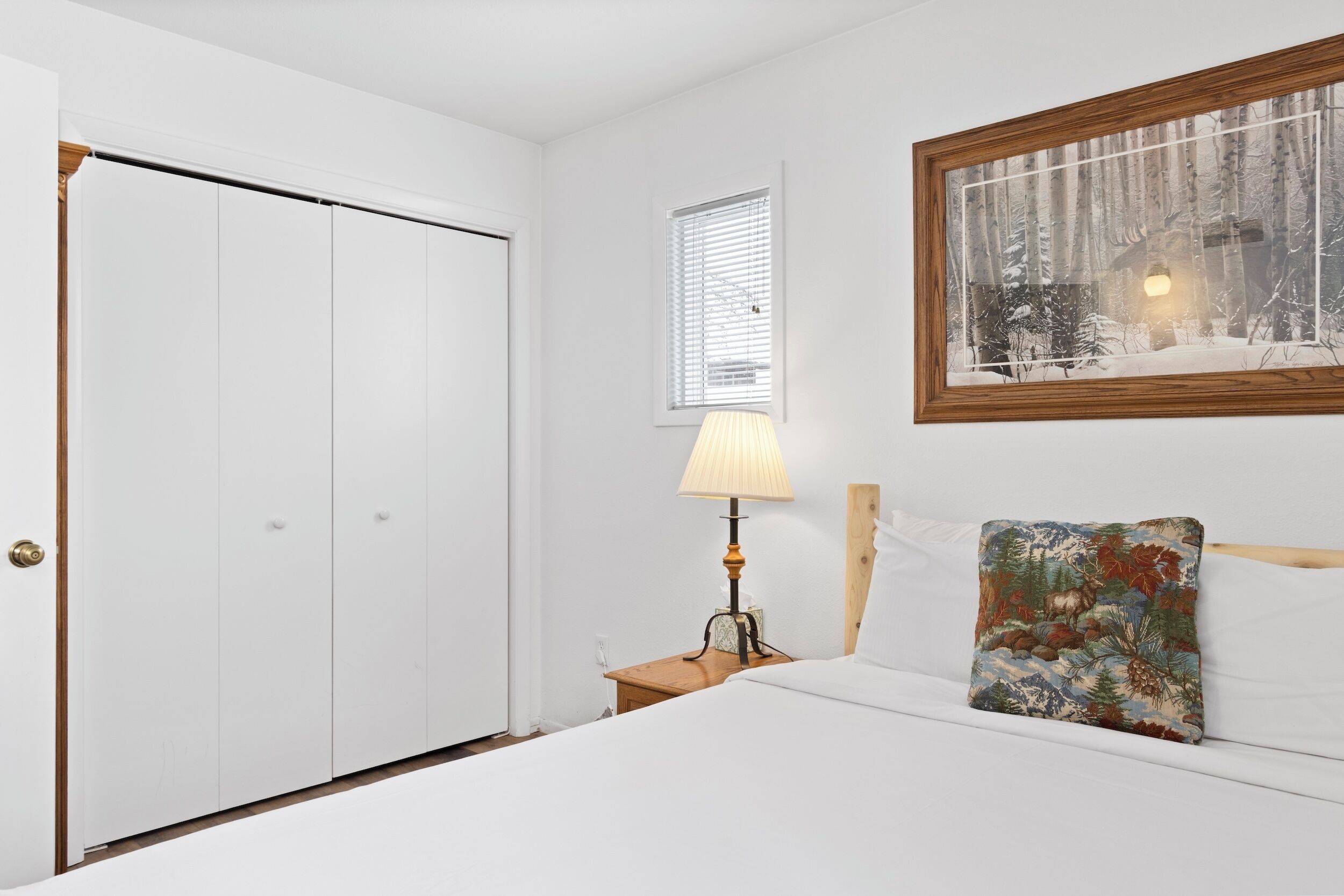 A bedroom with a bed, a nightstand with a lamp, a window with blinds, and a framed picture on the wall.