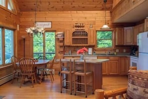 A wooden cabin interior with a dining area, kitchen, and large windows.