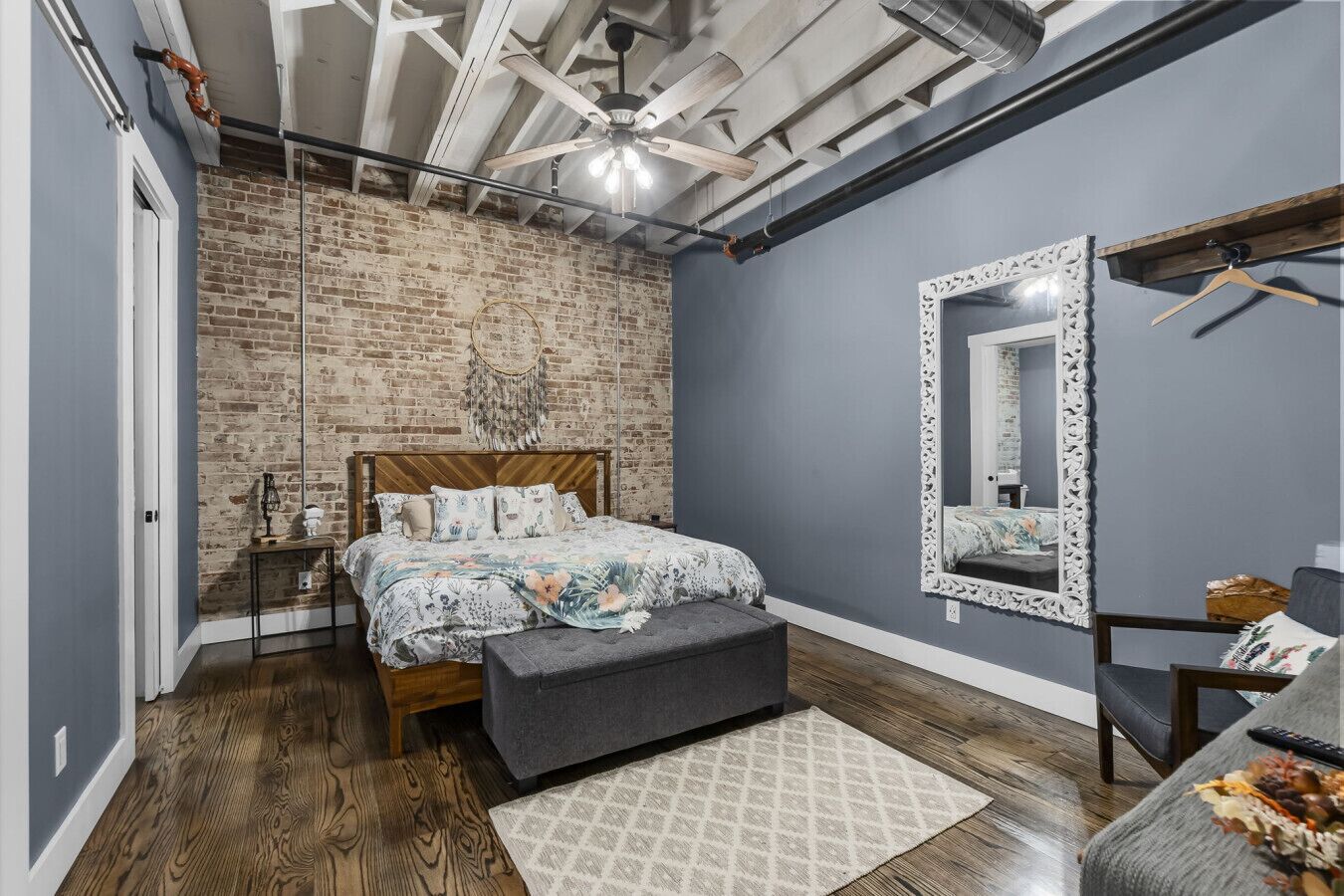 A bedroom with a brick wall, a bed with floral bedding, a mirror with an ornate frame, and a hanging light fixture.