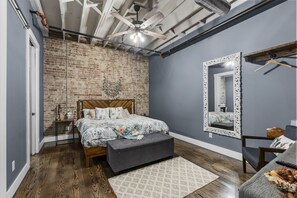 A bedroom with a brick wall, a bed with floral bedding, a grey bench, a mirror with an ornate frame, and a ceiling fan. - Urban Lofts (Perryville)