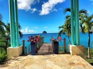 Un sentier dallé de pierres menant à une plage bordée de piliers turquoise et de fleurs roses.