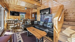 Ein gemütliches Blockhäuschen im Inneren mit Holzbalkendecke, einem schwarzen Ledersofa, einem Holztisch und einem an der Wand befestigten Fernseher.