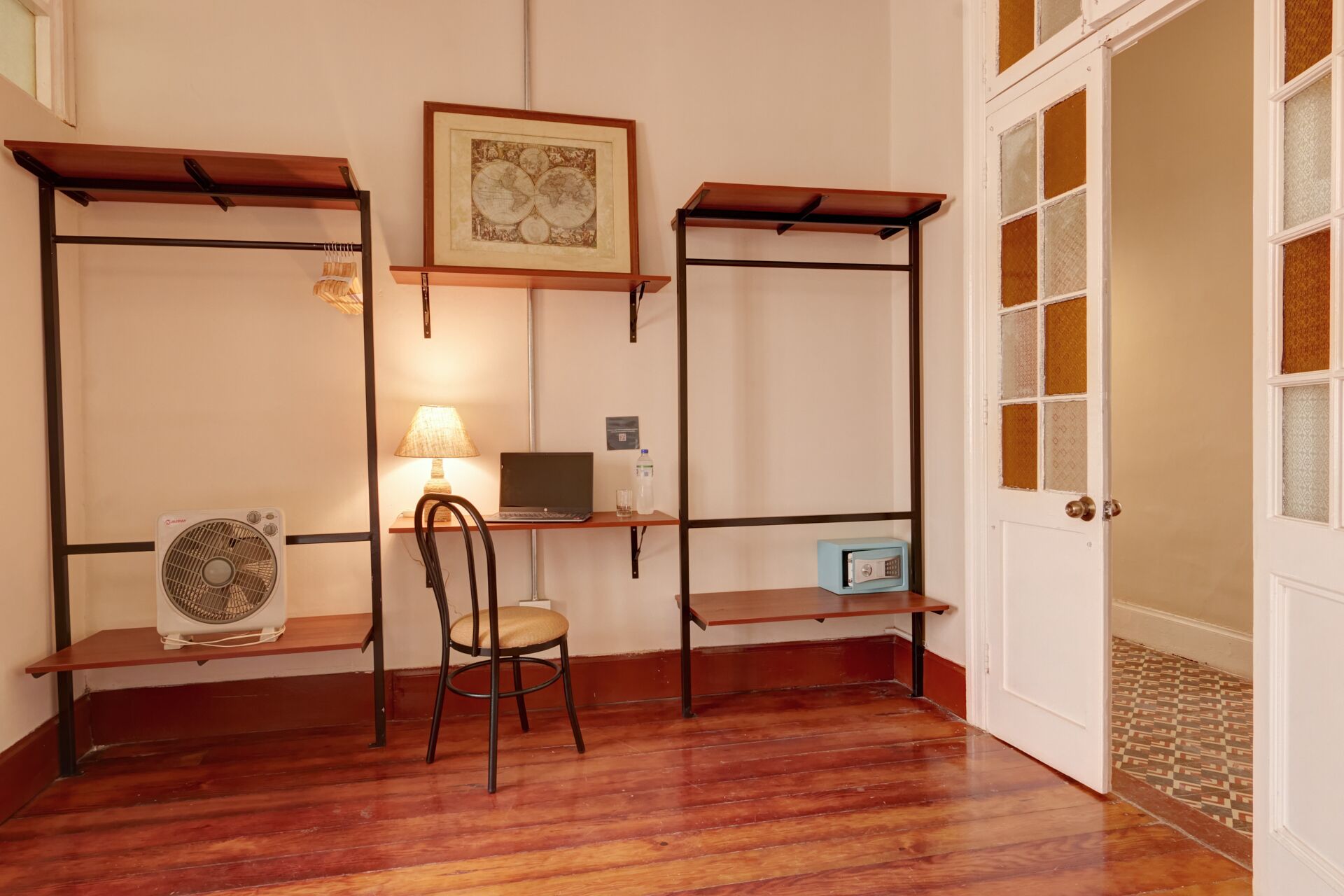 A room with a wooden floor, a desk with a laptop, a chair, a fan, and a shelf with a framed picture.