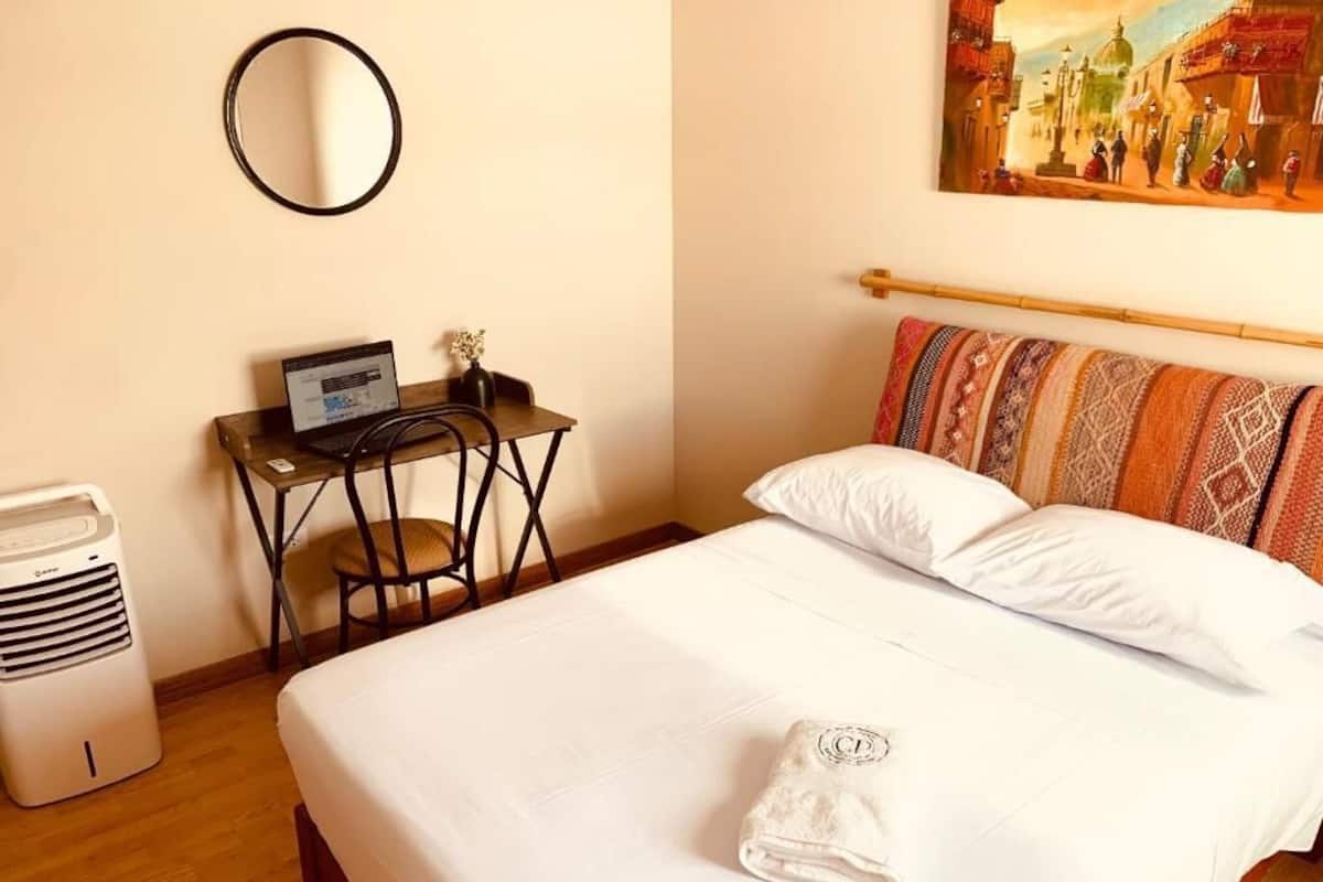 A neatly made bed with white linens, a laptop on a small table beside it, and a wall-mounted mirror.