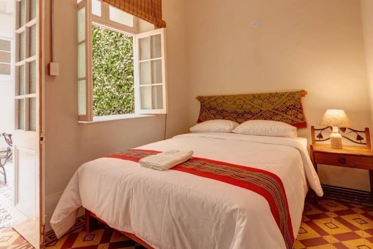 A neatly made bed with a white comforter and a red and yellow striped blanket, a wooden nightstand with a lamp, and a window with a view of greenery.
