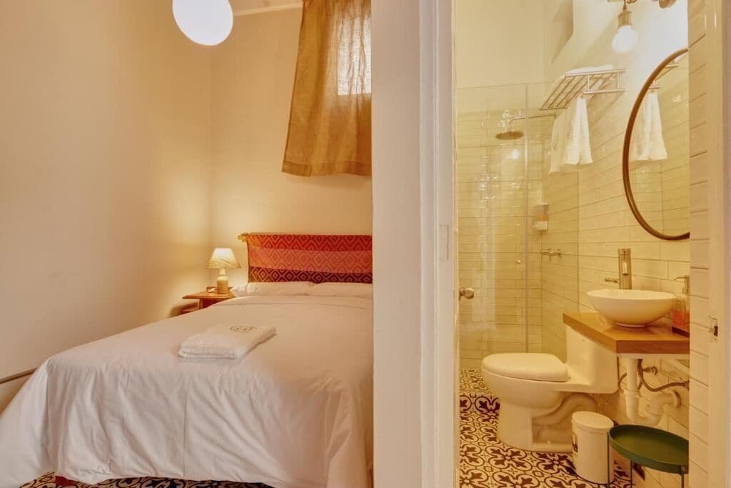 A hotel room with a bed, a bedside table with a lamp, a bathroom with a toilet, sink, and shower.