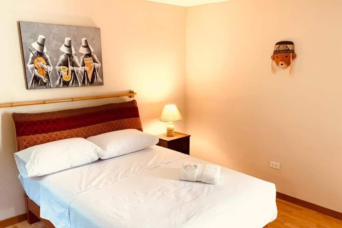 A neatly made bed with white linens, a headboard with a red and brown pattern, a wall-mounted lamp, and a painting of musicians.