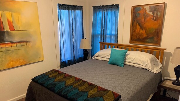 A neatly made bed with a patterned bedspread and pillows, a bedside table with a lamp, and a painting on the wall. - Casa Colorado: Cozy. Cute. Convenient. (Colorado Springs)