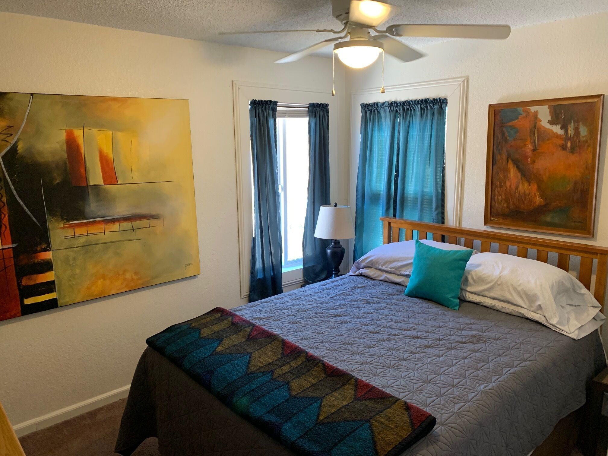 A hotel room with a bed, a ceiling fan, a painting on the wall, and a window with curtains.