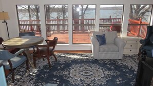 A room with a large window overlooking a wooden deck and a body of water. There is a round table with chairs, a striped armchair, and a small cabinet. - Eufaula Lake front access w/wraparound deck and spectacular views! Pet friendly! (Checotah)