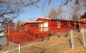 Une cabane rouge avec un balcon, une cour clôturée et un lac en arrière-plan.