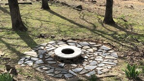A circular stone fire pit surrounded by a stone border, set in a grassy area with trees in the background. - Eufaula Lake front access w/wraparound deck and spectacular views! Pet friendly! (Checotah)