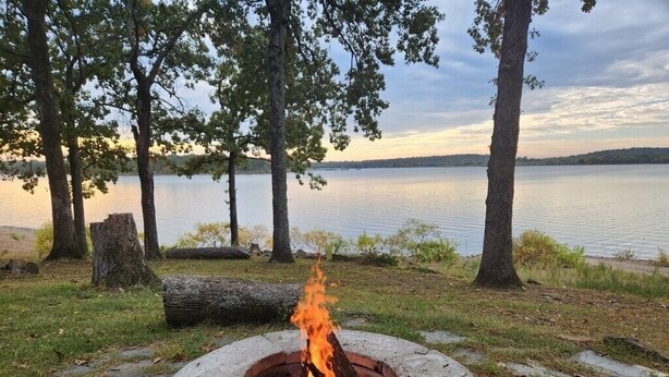 A fire pit in a natural setting with a lake and trees in the background. - Eufaula Lake front access w/wraparound deck and spectacular views! Pet friendly! (Checotah)