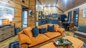 A cozy cabin interior with a wooden ceiling, a sofa with cushions, a dining table with chairs, and a kitchen area. - Romantic|Private|Wellness|OutdoorMovieNight (Broken Bow)
