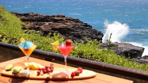 A table with drinks and fruit, overlooking a rocky coastline and ocean waves. - Oceanfront Condo with Stunning Views & AC (Koloa)