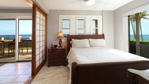 A bedroom with a bed, a nightstand, a lamp, and a view of the ocean through large windows. - Oceanfront Condo with Stunning Views & AC (Koloa)