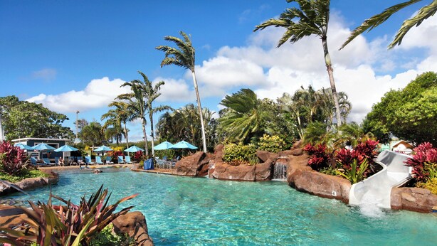 A clear pool with a water slide, surrounded by palm trees and lounge chairs. - Oceanfront Condo with Stunning Views & AC (Koloa)