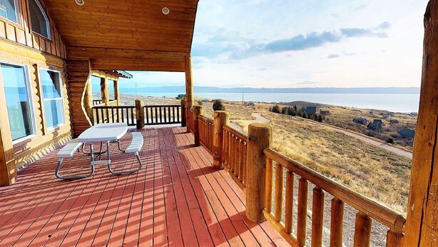 Une terrasse en bois avec une table et des chaises, offrant une vue panoramique sur un paysage pittoresque comprenant un plan d’eau et des collines.