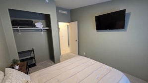 A bedroom with a bed, a TV mounted on the wall, a closet with a towel rack, and a door leading to another room.