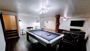 A pool table with billiard balls, a television, and a wooden tree trunk in a room with white walls and wooden flooring.