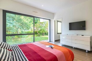 A modern bedroom with a large sliding glass door leading to a garden, a bed with striped bedding, a white dresser, and a flat-screen TV mounted on the wall.