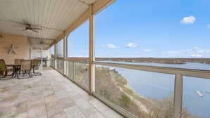Une terrasse couverte avec une table et des chaises, de grandes fenêtres donnant sur un plan d’eau et un paysage lointain.