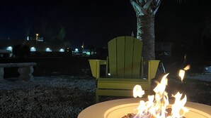 A circular outdoor fire pit with visible flames, surrounded by gravel, with a yellow chair and a palm tree in the background.