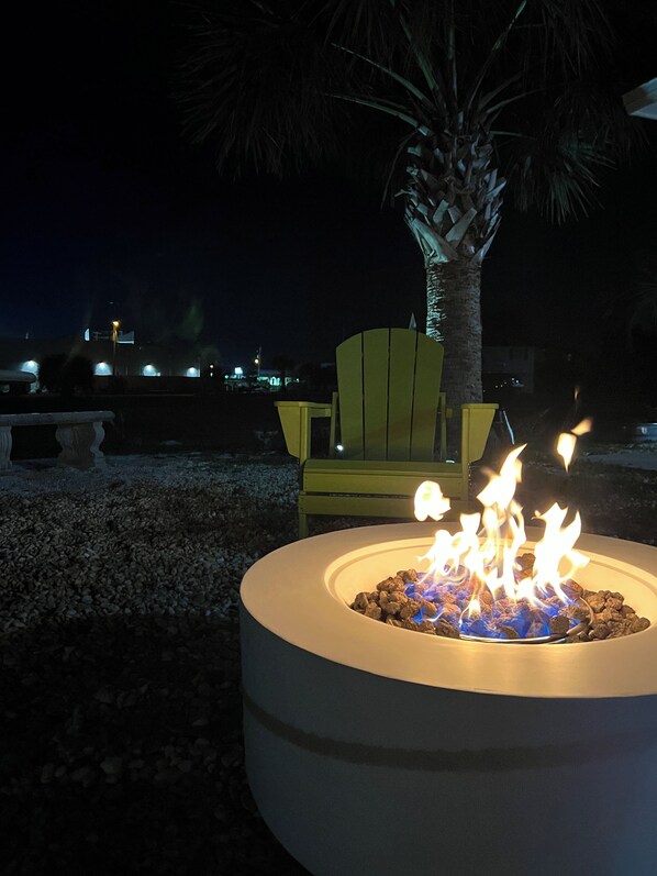 A circular outdoor fire pit with visible flames, surrounded by gravel, with a yellow chair and a palm tree in the background. - Fully Renovated, Steps from Beach with Gulf Views. Pet Friendly! (Panama City Beach)