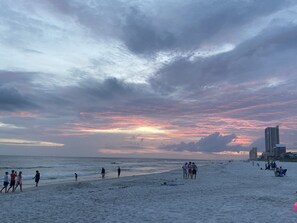 A beach with people walking and standing, and buildings in the background.