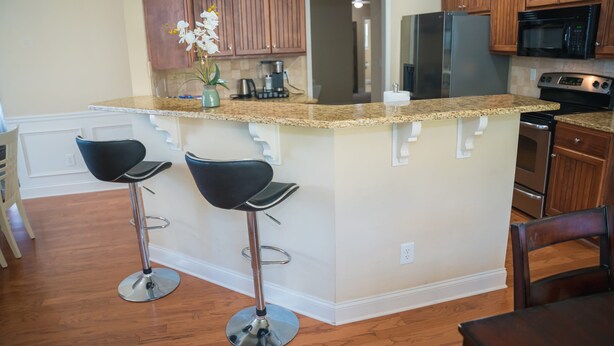 A kitchen with wooden cabinets, a granite countertop, stainless steel appliances, and two black bar stools. - Craftsman ranch (Lawrenceville)