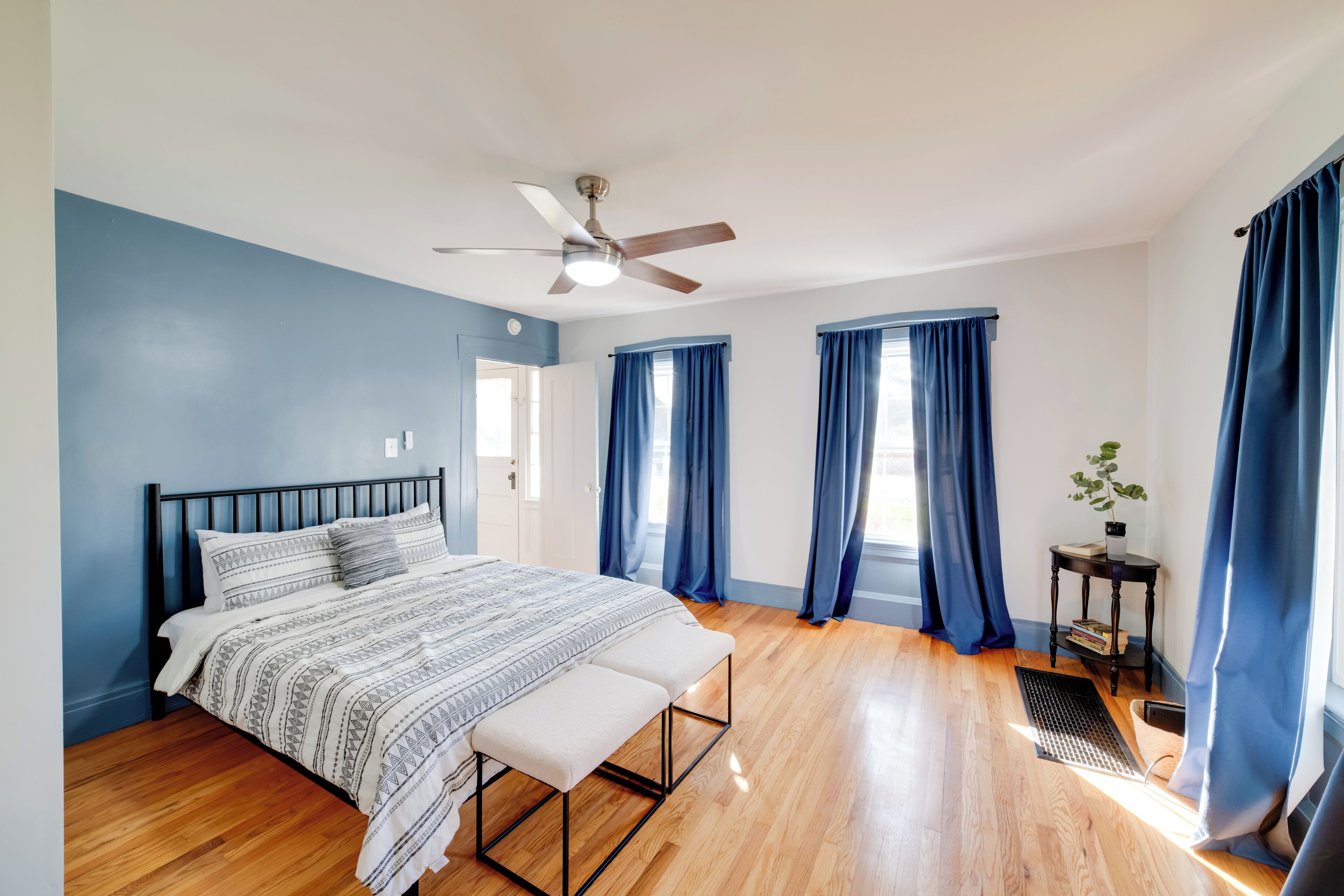 Un dormitorio con cama, un banco, ventilador de techo y una pequeña mesa con planta.