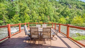 Een houten terras met een tafel en stoelen, met uitzicht op een rivier en bebost gebied.